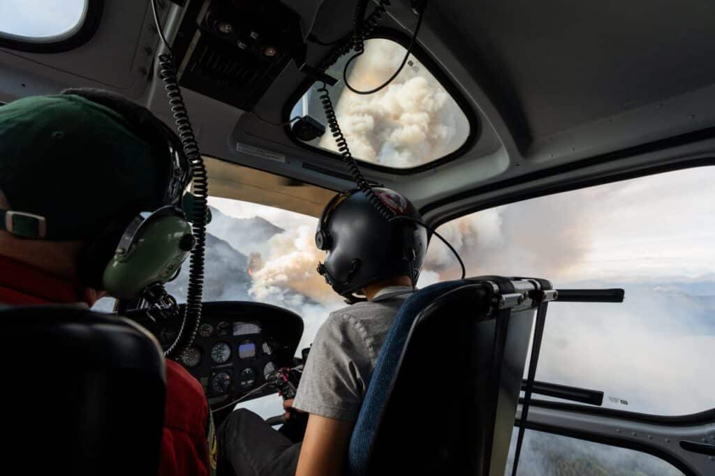 A view from inside a helicopter and over the shoulders of a pair of pilots shows a roiling cloud of smoke heaving up from an orange flame on a mountain slope.