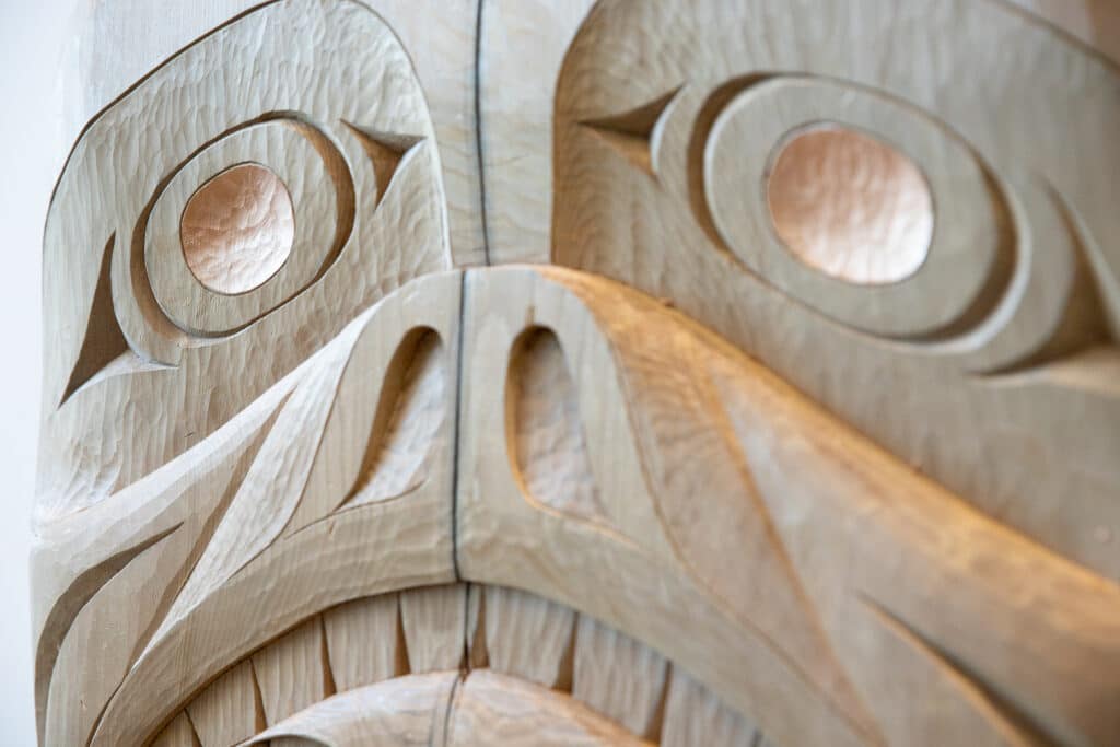 A close-up of the face of a bear with copper-leafed eyes carved in Squamish style into a wooden house post.