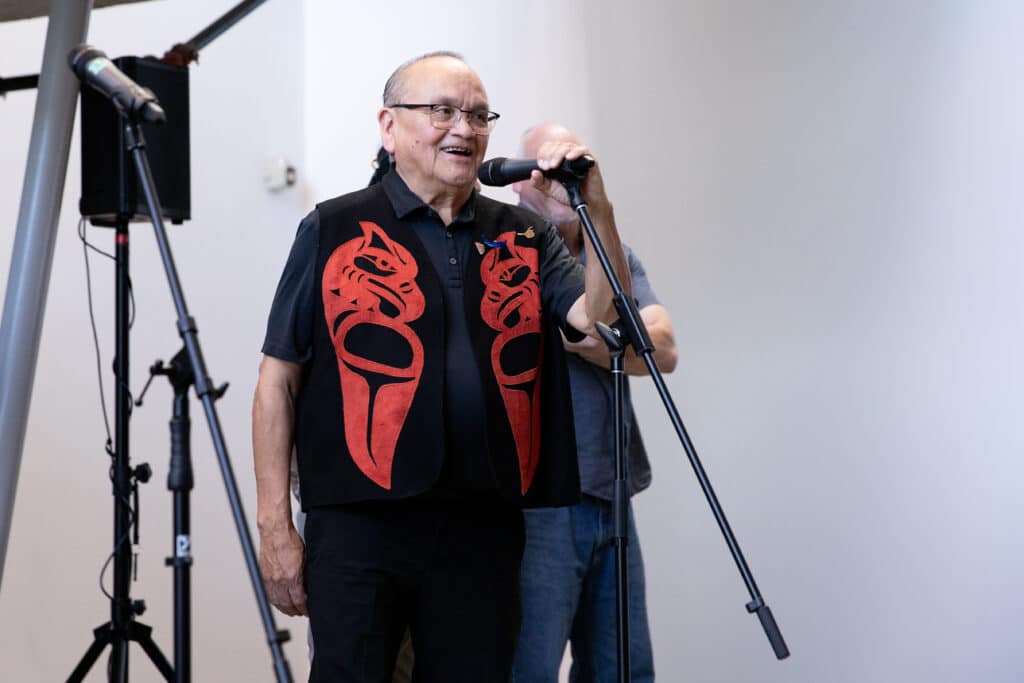 A person in a black vest with red Coast Salish designs stands smiling at a microphone.