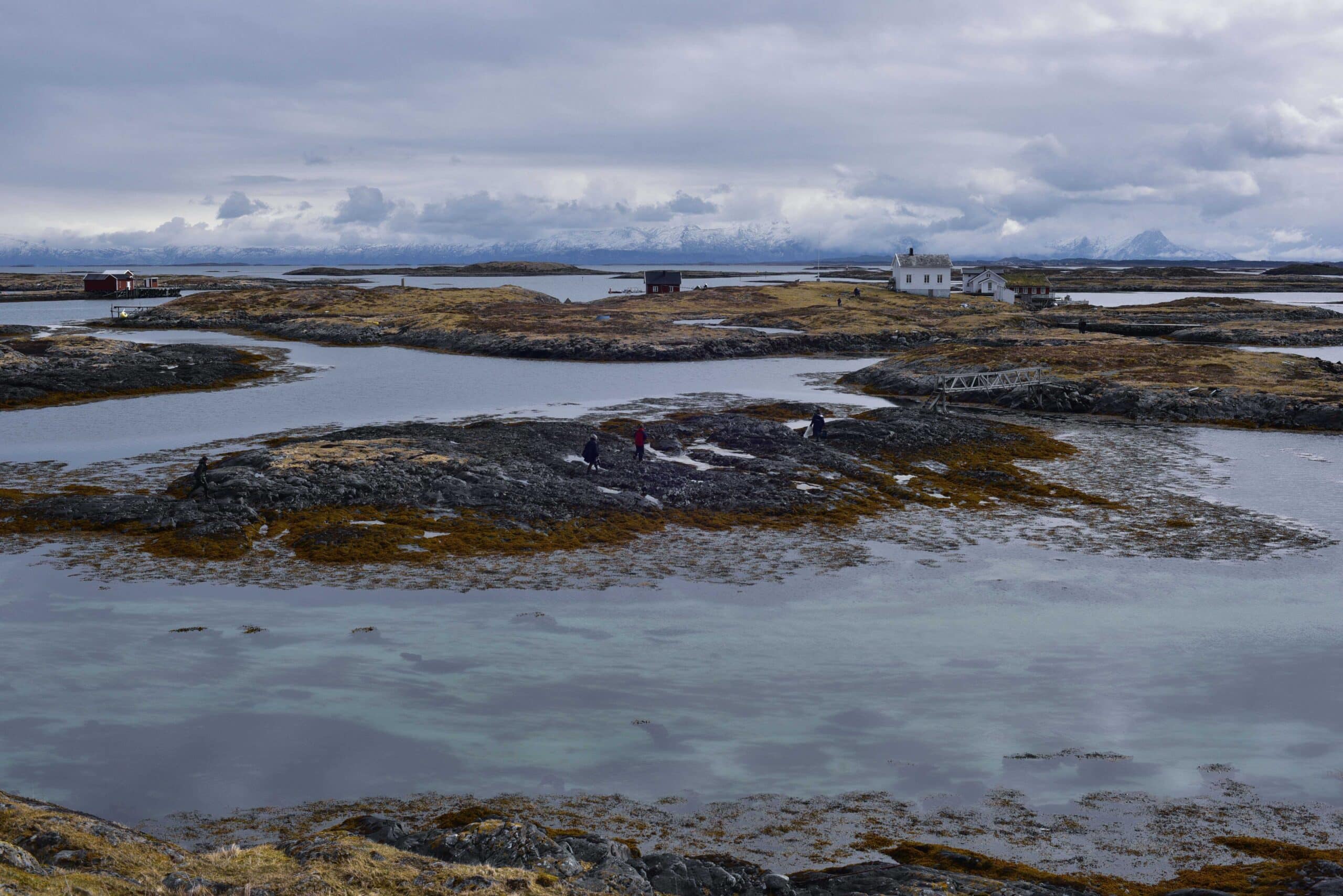 A wide coastal landscape showing rocky islands, shallow water, and scattered buildings under a cloudy sky, with several small figures walking along the shore.