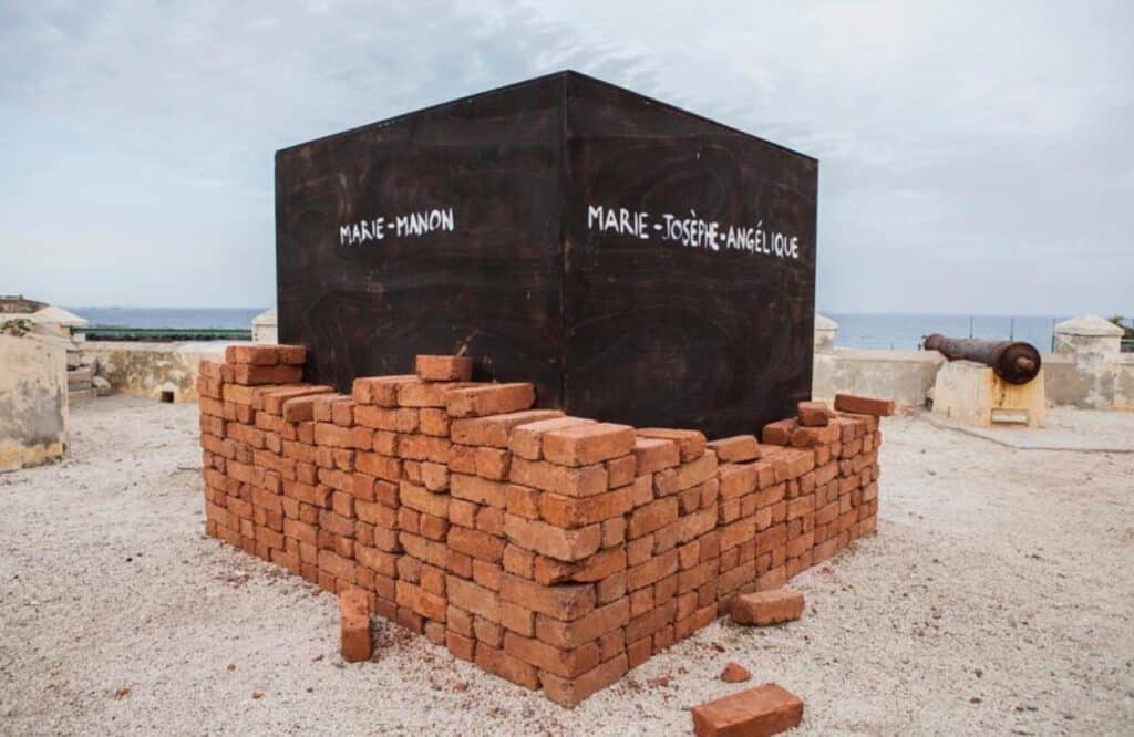 Outdoor installation of a black cube surrounded by stacked red bricks, set near the ocean.