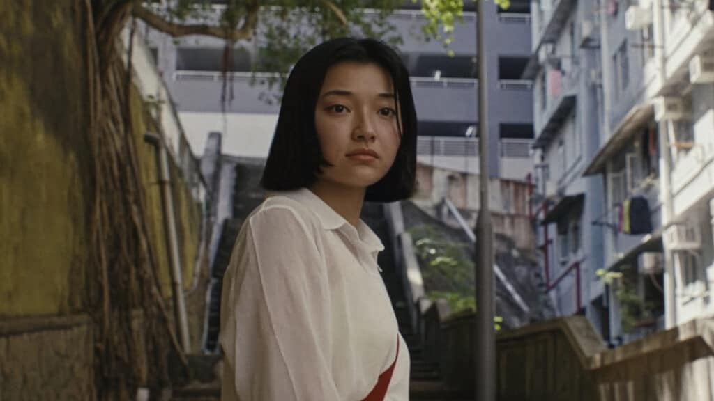 A person with a short haircut stands on an outdoor stairway between apartment buildings, looking toward the camera as daylight filters through nearby trees.