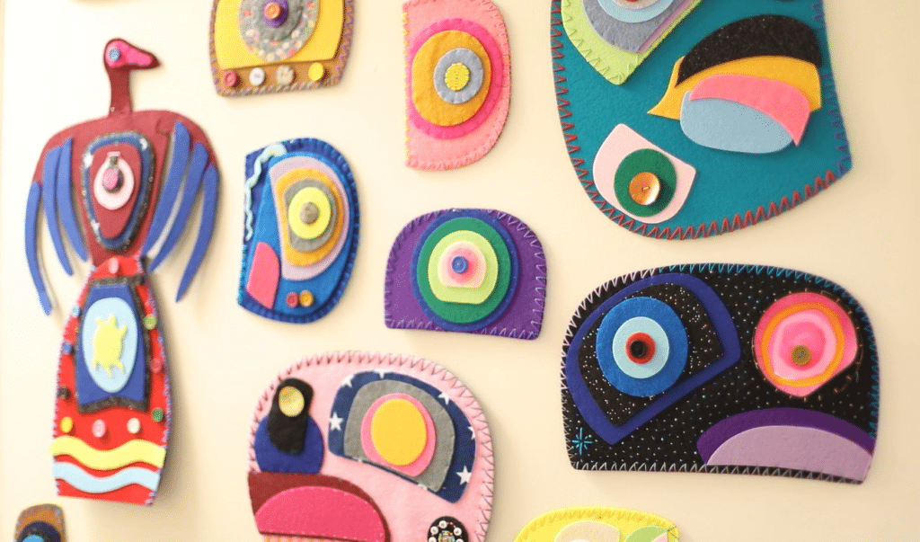 Colourful handmade textile artworks with layered shapes and stitched details arranged on a wall, including a bird figure and abstract circular designs.