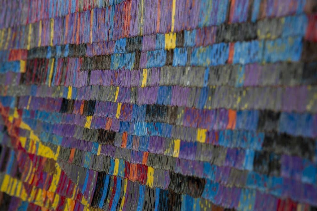 Close-up of a woven surface made from short, dense strands of colour arranged in horizontal bands of purple, blue, yellow and charcoal, creating a rhythmic, tactile pattern.