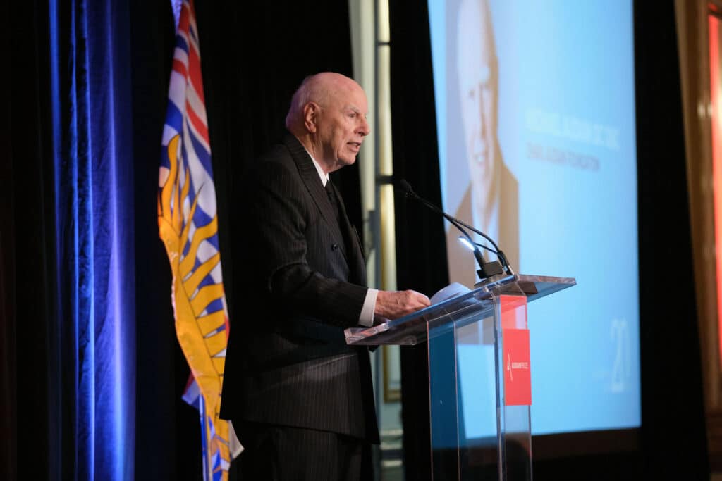 An individual stands at a clear podium delivering a speech, framed by stage lighting and a large projected image behind them, with a provincial flag visible to one side.