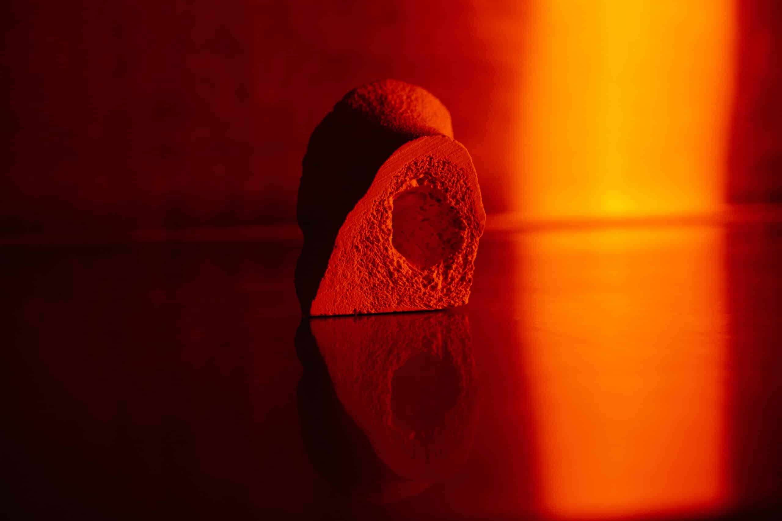 An abstract, stone-like form rests on a reflective surface, illuminated by deep red and orange light. Its textured surface and rounded hollow are mirrored below, creating a quiet, glowing composition.