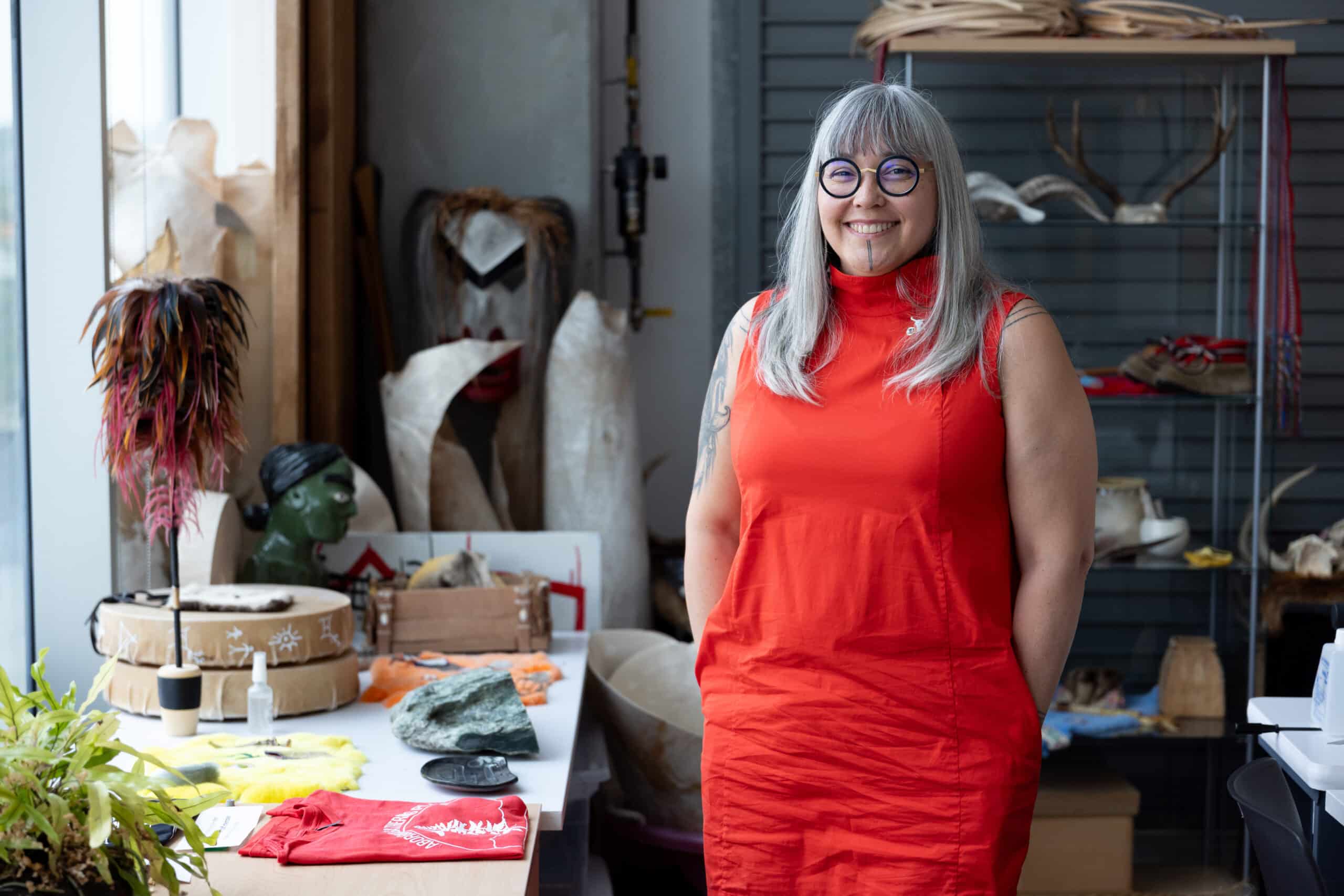 A person stands smiling in a studio filled with natural materials and sculptural forms, wearing a bright red garment amid shelves, tools and works in progress.