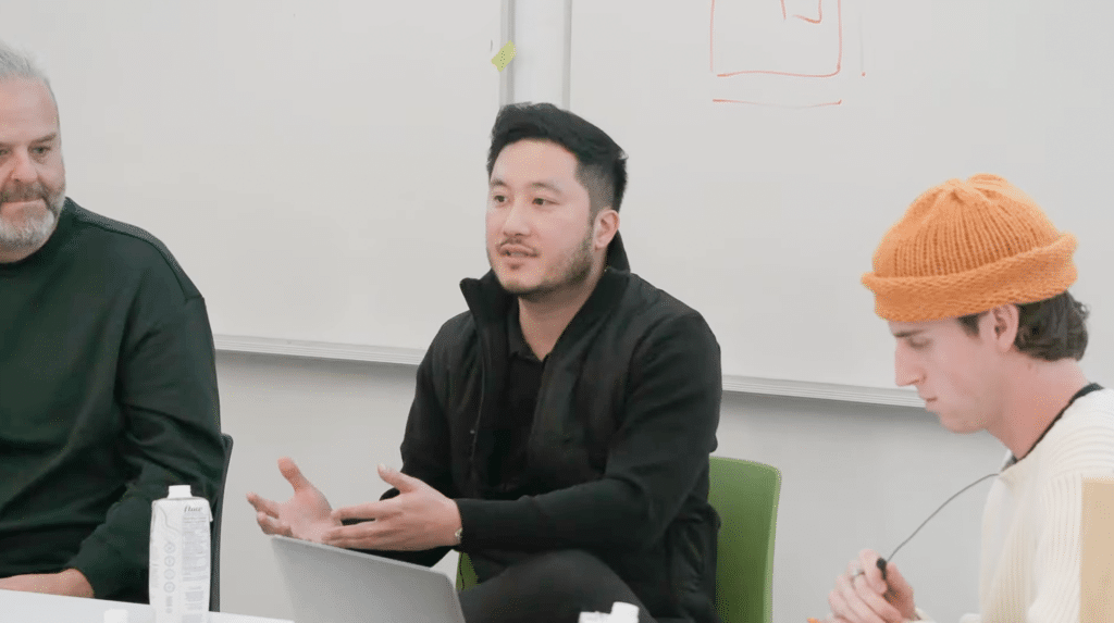 People sit together at a table in a classroom setting, engaged in discussion. One person gestures while speaking, with a laptop, notebooks and drinks on the table and a whiteboard in the background.