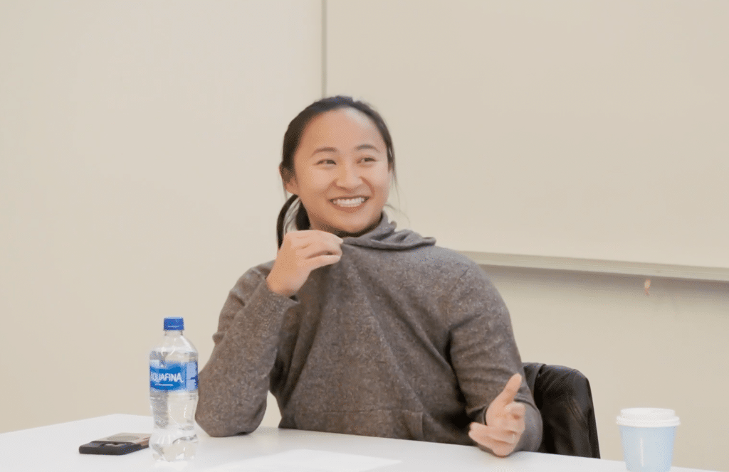 A person sits at a table in a classroom setting, smiling mid-conversation. A water bottle and coffee cup rest nearby, with a whiteboard behind them and a relaxed, conversational moment unfolding.
