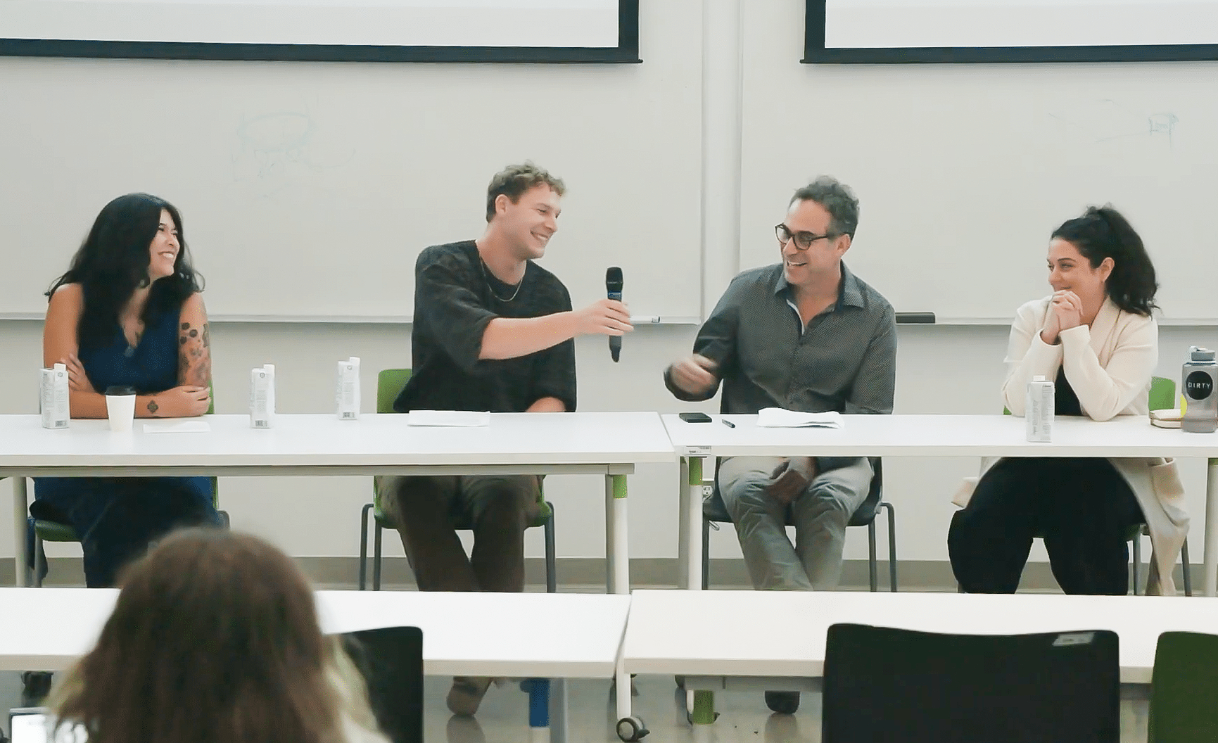 Four people sit at a table in a classroom setting, smiling and engaged in conversation. One person passes a microphone along the table as others listen, with notebooks, drinks and a whiteboard visible behind them.