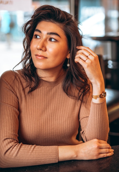 A person seated at a table looks thoughtfully to the side, one hand brushing their hair, with warm indoor light softening their expression and the background gently out of focus.