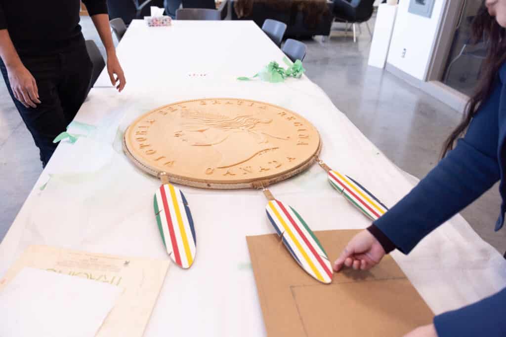 A large carved wooden medallion rests on a table, its surface etched with a profile and circular text, three striped feather forms hanging below. Hands nearby carefully position materials around it, suggesting a moment of preparation and care.