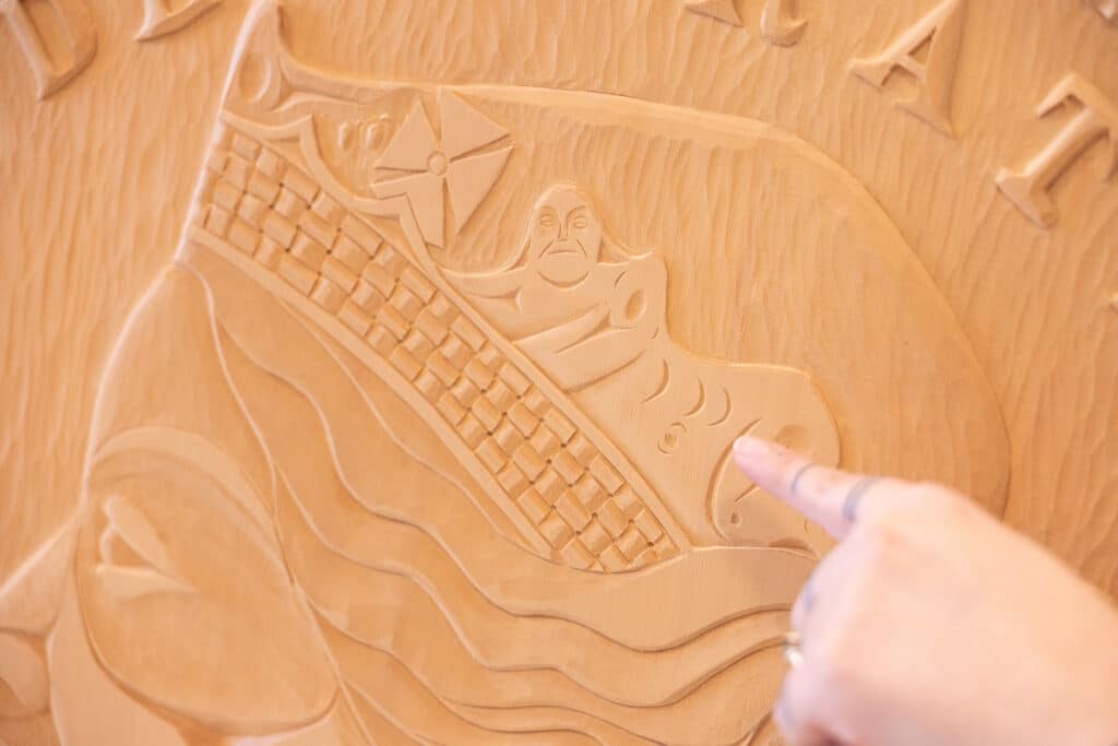Close-up of a carved wooden medallion, showing a detailed relief of a figure beside flowing lines and woven patterns. A hand gently points to part of the carving, drawing attention to the fine craftsmanship in the pale wood grain.