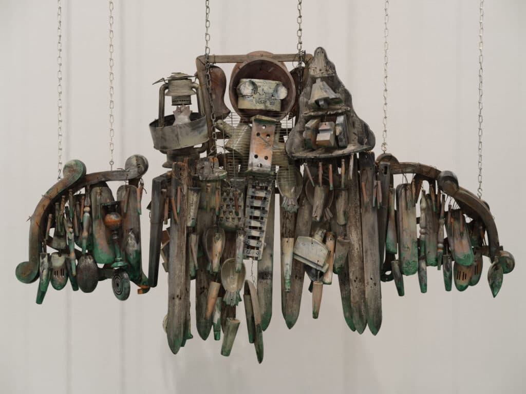 A suspended sculpture made from weathered wood and metal tools hangs against a white wall, its shape suggesting outstretched wings or a mechanical figure.