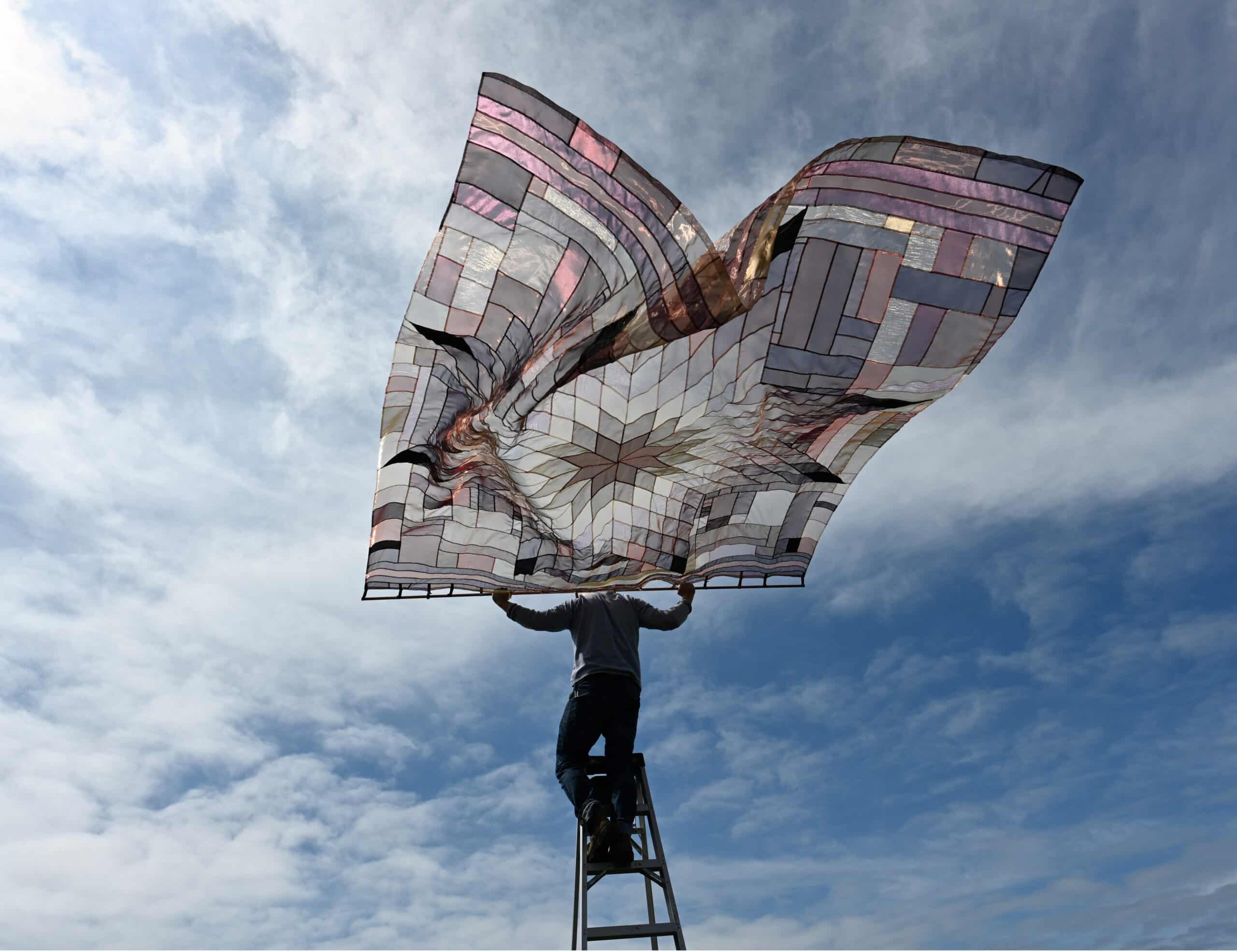 A person stands on a ladder outdoors holding up a large patterned textile that billows against a cloudy sky