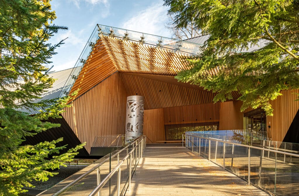 The entrance to a contemporary gallery clad in warm wood, where angled beams and glass railings lead visitors along a ramp framed by evergreen trees and soft afternoon light.
