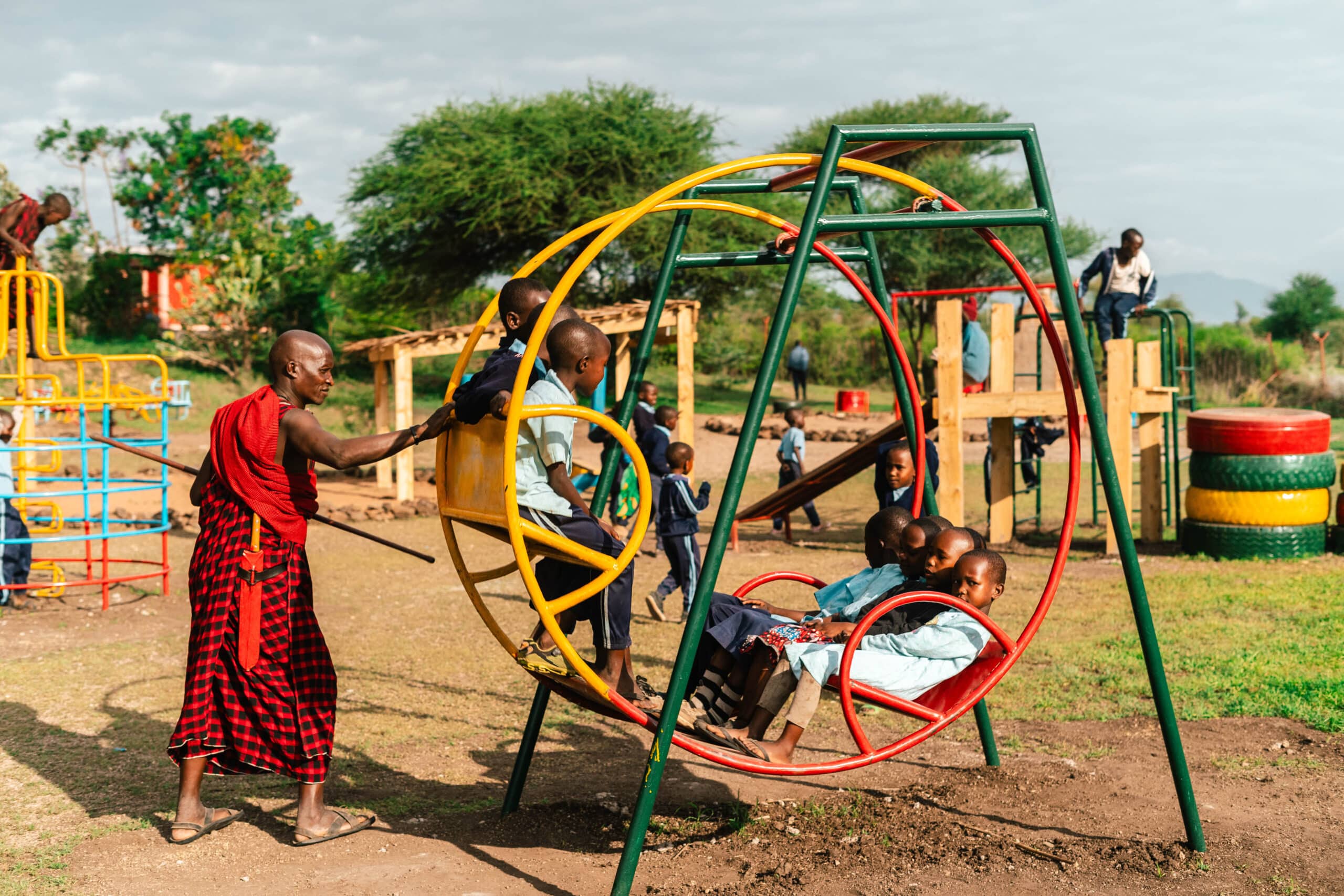 Children ride a circular metal swing in an open playground as an adult gives a careful push. Brightly painted play structures sit among grass and trees under an open sky.