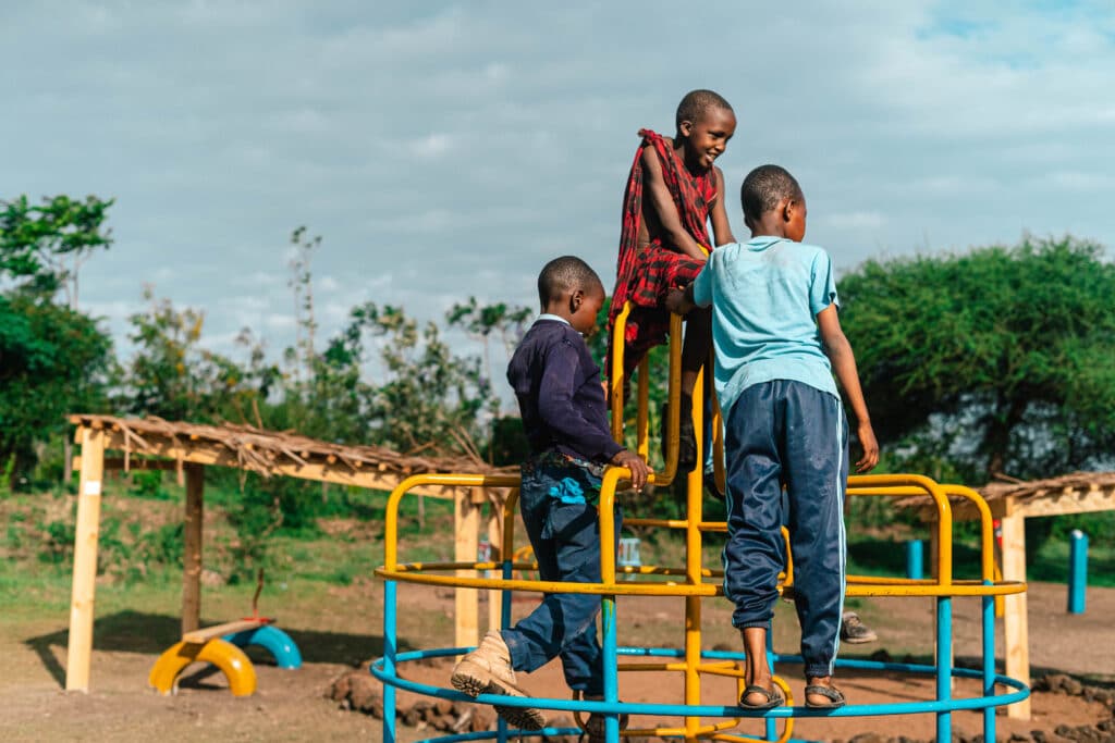 Children climb and balance on a circular metal play structure in an outdoor playground. Yellow and blue bars frame their movement as trees, playground equipment and open ground stretch into the background under a wide blue sky.
