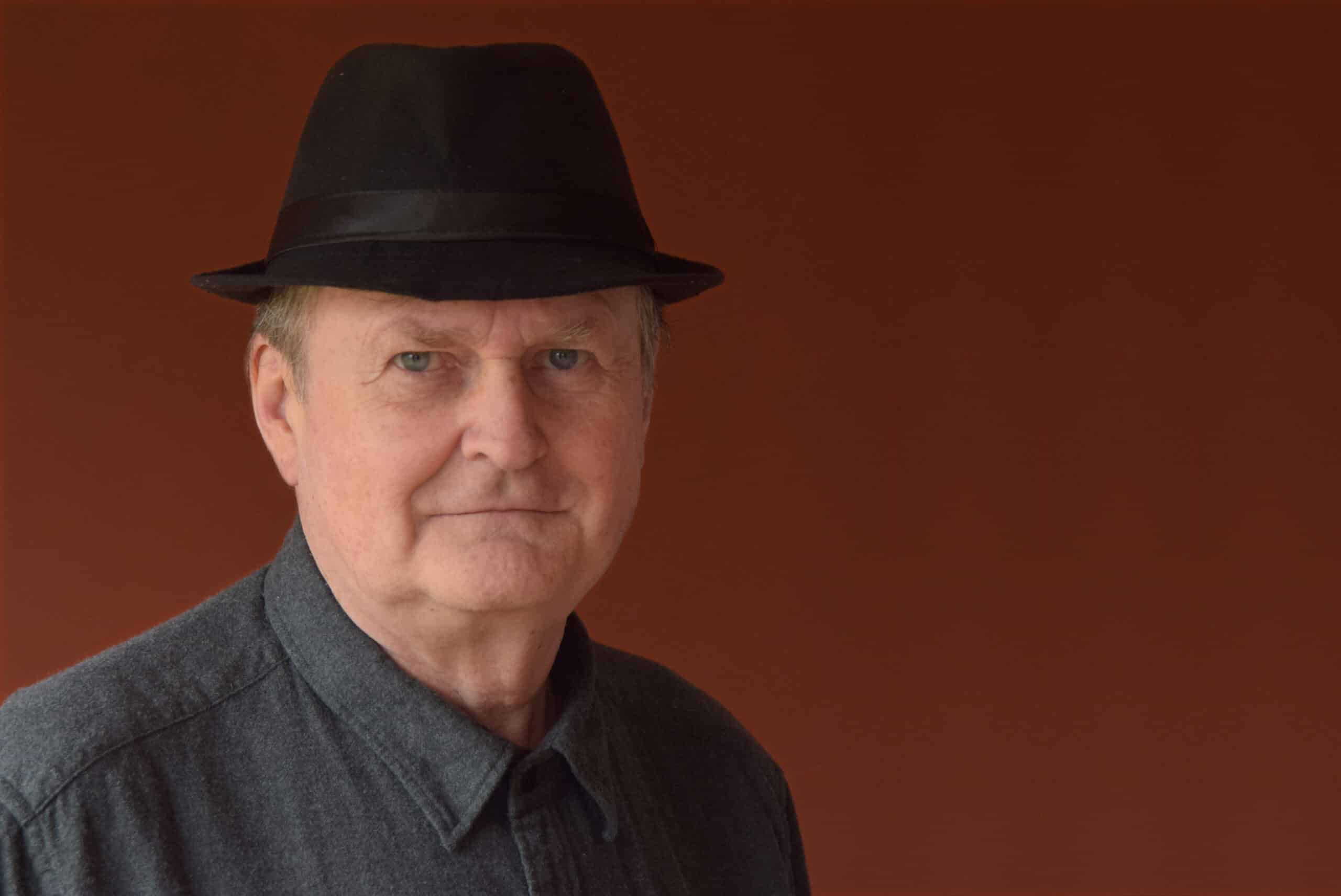 A person wearing a dark hat and buttoned shirt faces the camera against a warm, gradient background
