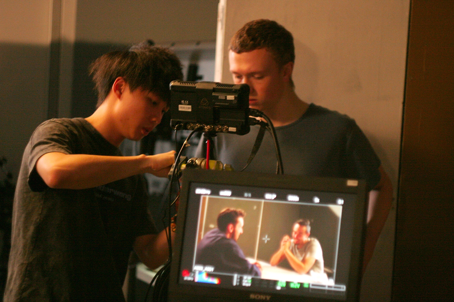 Two students collaborate in a dimly lit film production space, with one adjusting a professional video camera rig while the other observes a Sony monitor displaying a scene of two men conversing at a table. The setup highlights technical precision and teamwork in a hands-on studio environment.