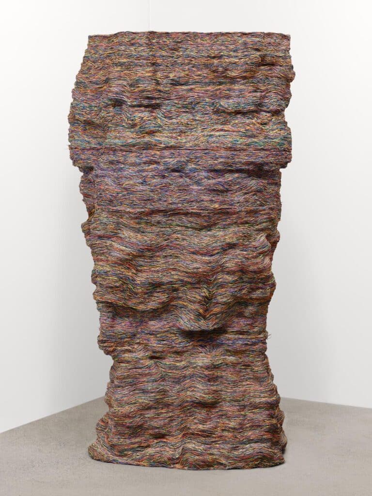 A tall, rectangular sculpture built from tightly layered, multicoloured threads, stacked in soft horizontal waves and standing upright against a white gallery wall.