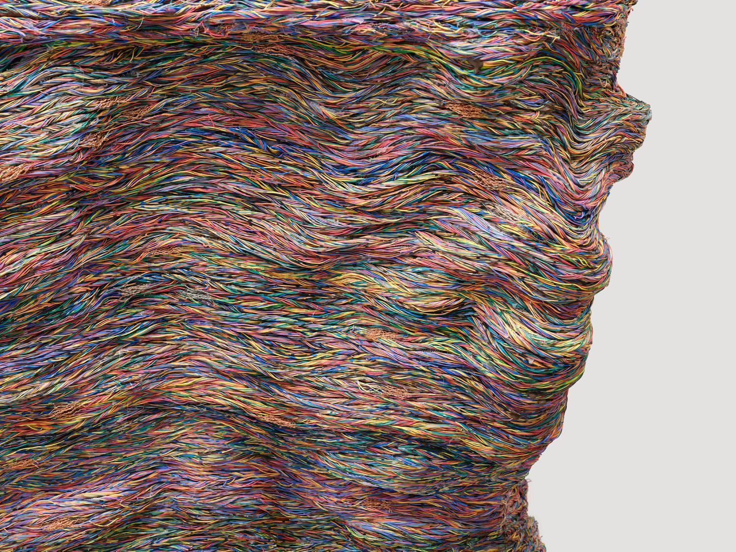 Close-up of a sculptural surface formed from flowing, multicoloured cords, compressed into rippling layers that suggest movement and weight.