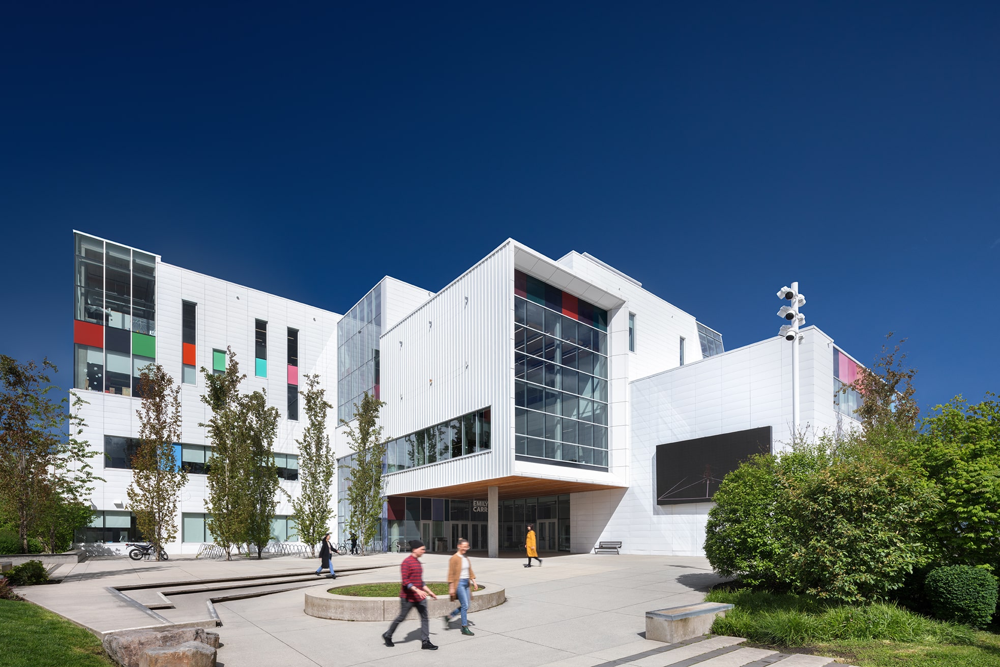 A modern white building opens onto a broad plaza under a deep blue sky. Glass walls and colorful window panels catch the light as people walk across the open space, framed by trees and landscaped paths.