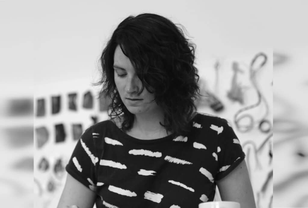A person with shoulder-length hair looks downward while working in a studio, surrounded by blurred hanging materials and tools in the background.