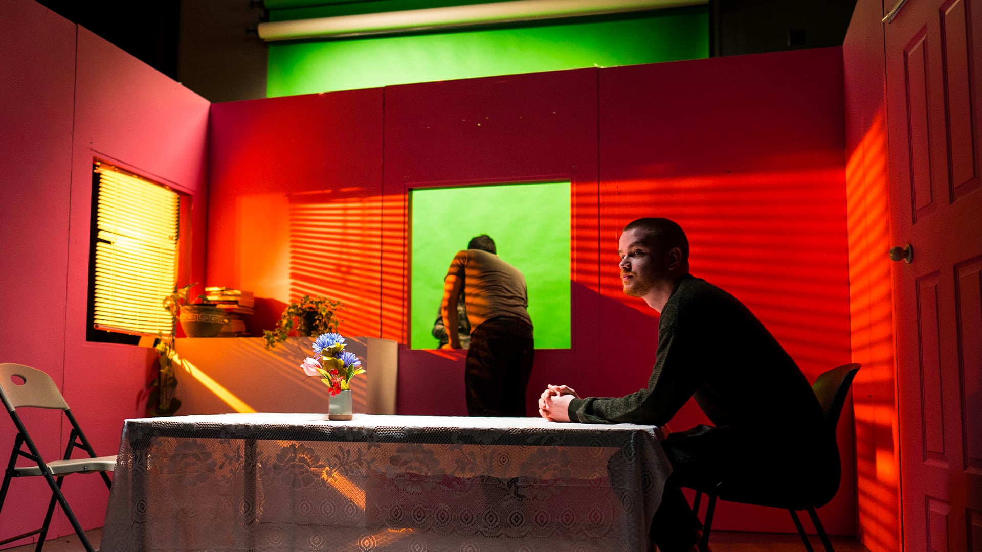 A person sits at a table covered with a lace tablecloth and a small vase of colorful flowers in a studio with vibrant pink walls. Another individual adjusts equipment in front of a green screen, while sunlight streams through window blinds, casting striped shadows. The space includes potted plants, a folding chair, and a closed door.