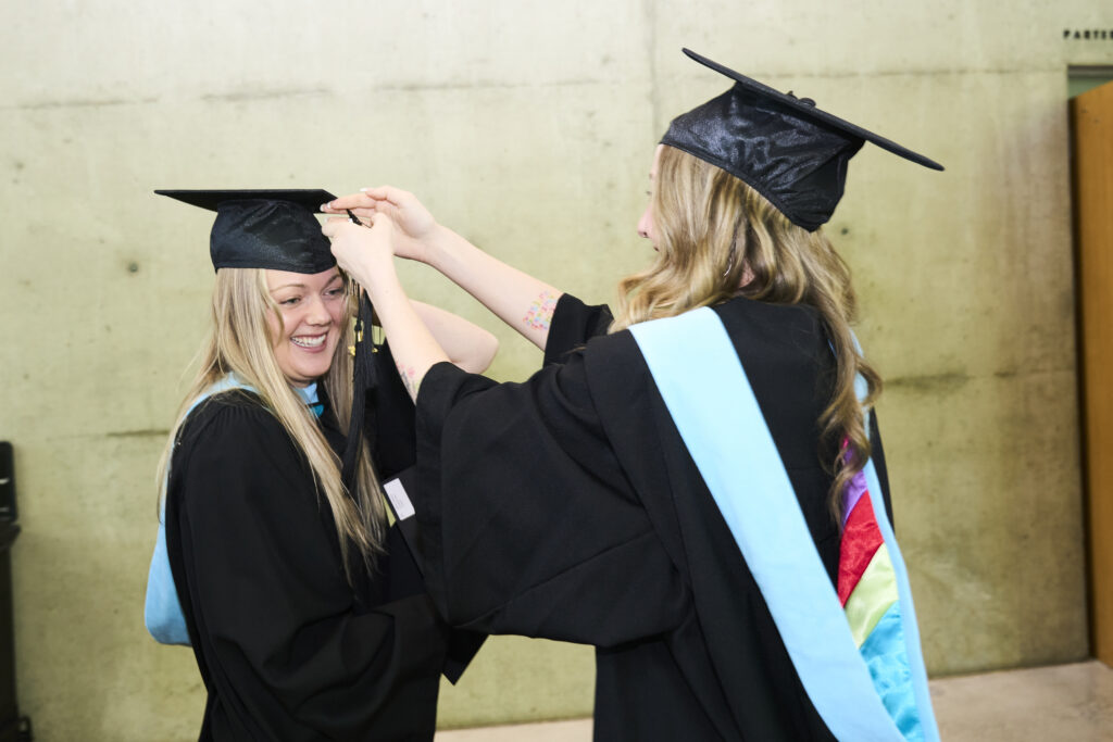 Two graduates in black gowns smile as one adjusts the other’s mortarboard tassel before the ceremony.