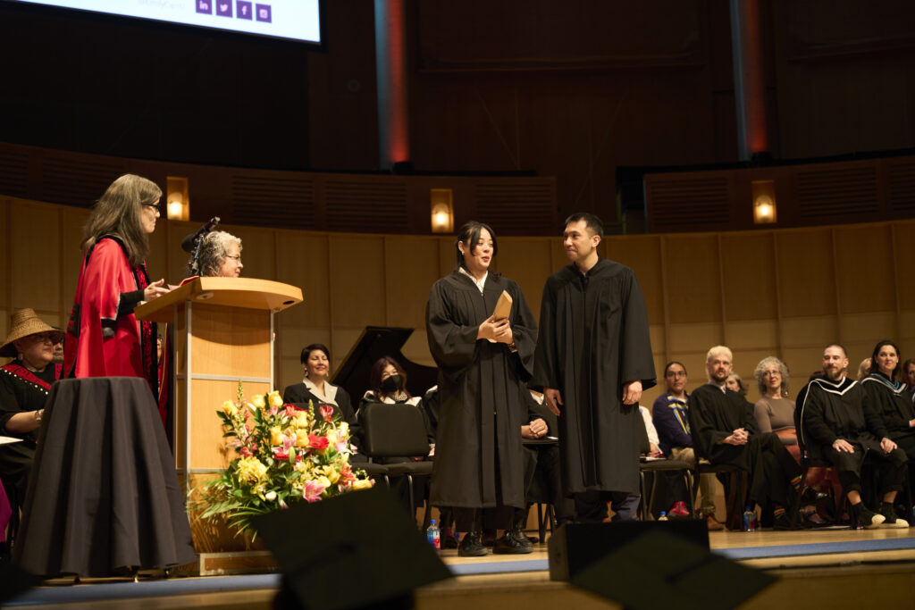Two people in black gowns stand on stage holding a folder while faculty look on during the convocation ceremony.