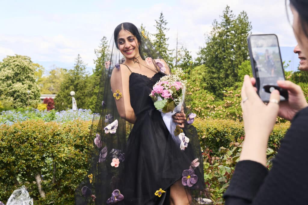 A graduate in a black dress with a sheer floral veil smiles while holding a bouquet as someone photographs her outdoors.