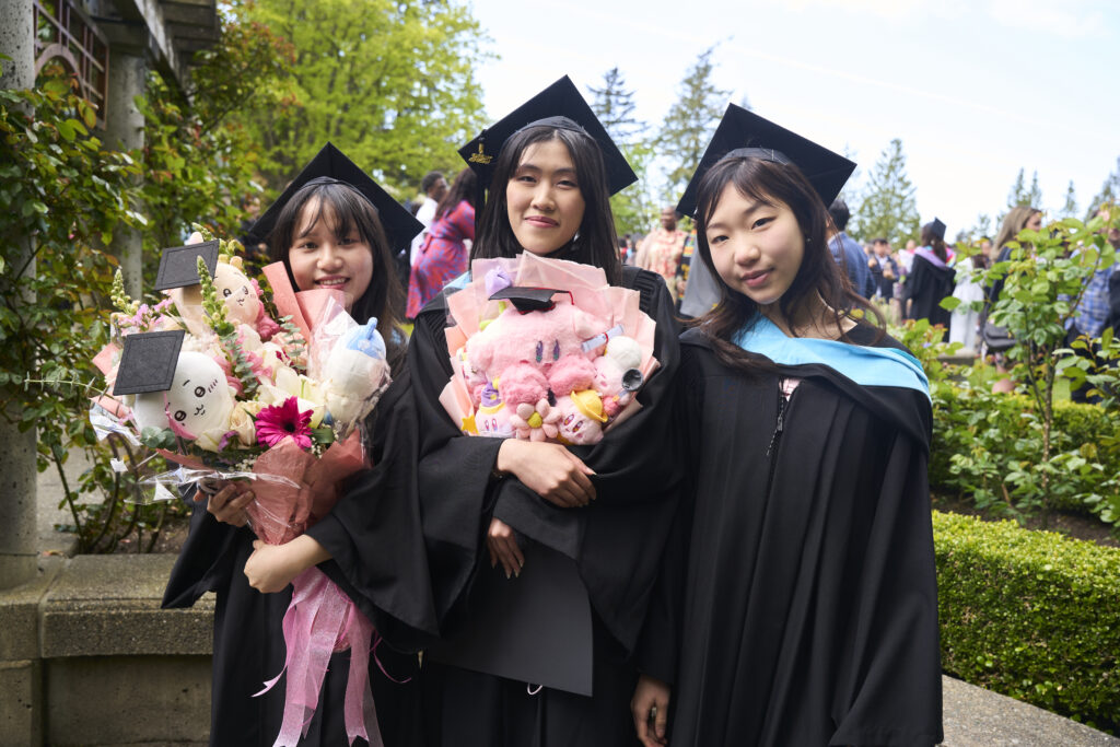 Three graduates in caps and gowns pose outdoors holding bouquets and plush gifts, smiling at the camera.