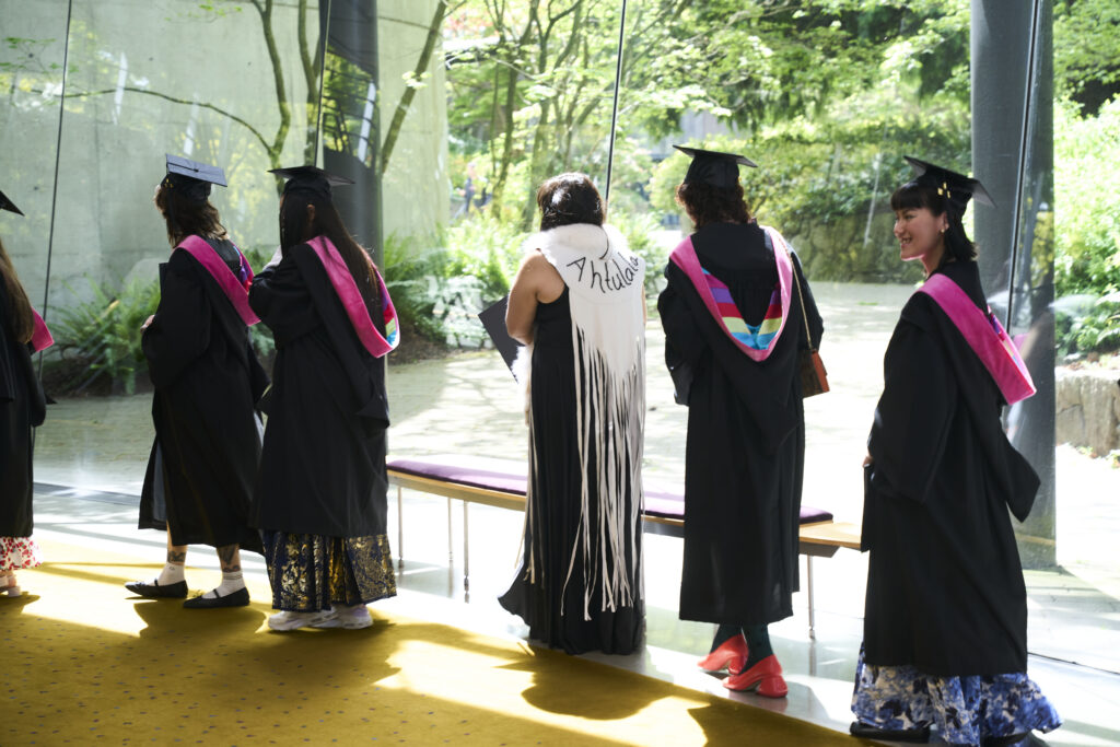 Graduates in caps and gowns stand in line near a large window, sunlight streaming in as they prepare for the ceremony.