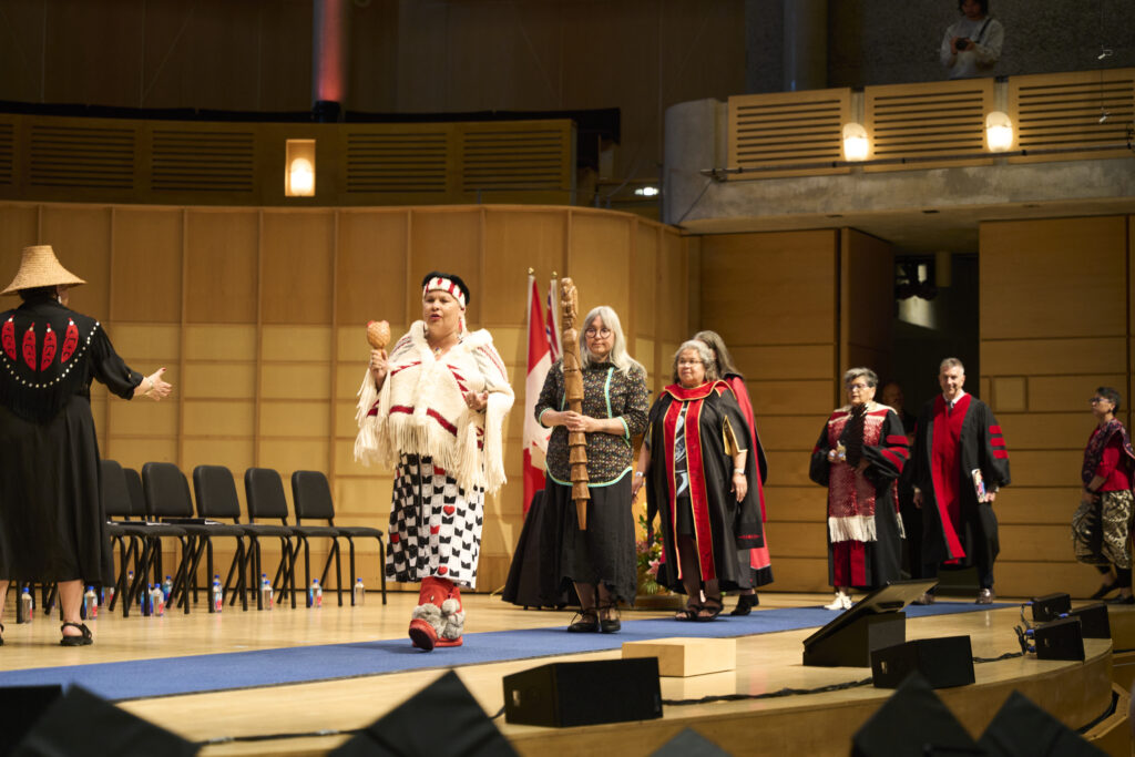 Indigenous leaders and academic officials process across a convocation stage in ceremonial regalia, with flags and faculty seated behind them.