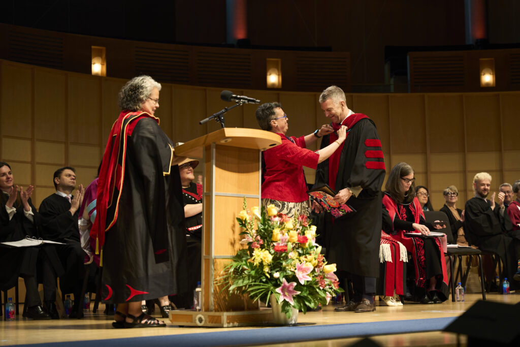 An academic official places a red hood over a man’s shoulders on stage as faculty applaud.