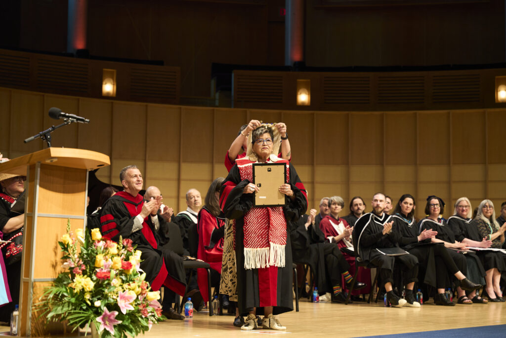 A woman in ceremonial dress holds a framed certificate while another person places a medallion over her head during the ceremony.