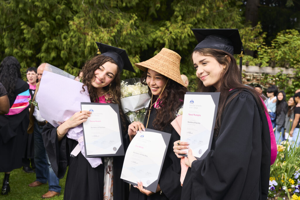 Three graduates in caps and gowns pose outdoors holding their diplomas and flowers.