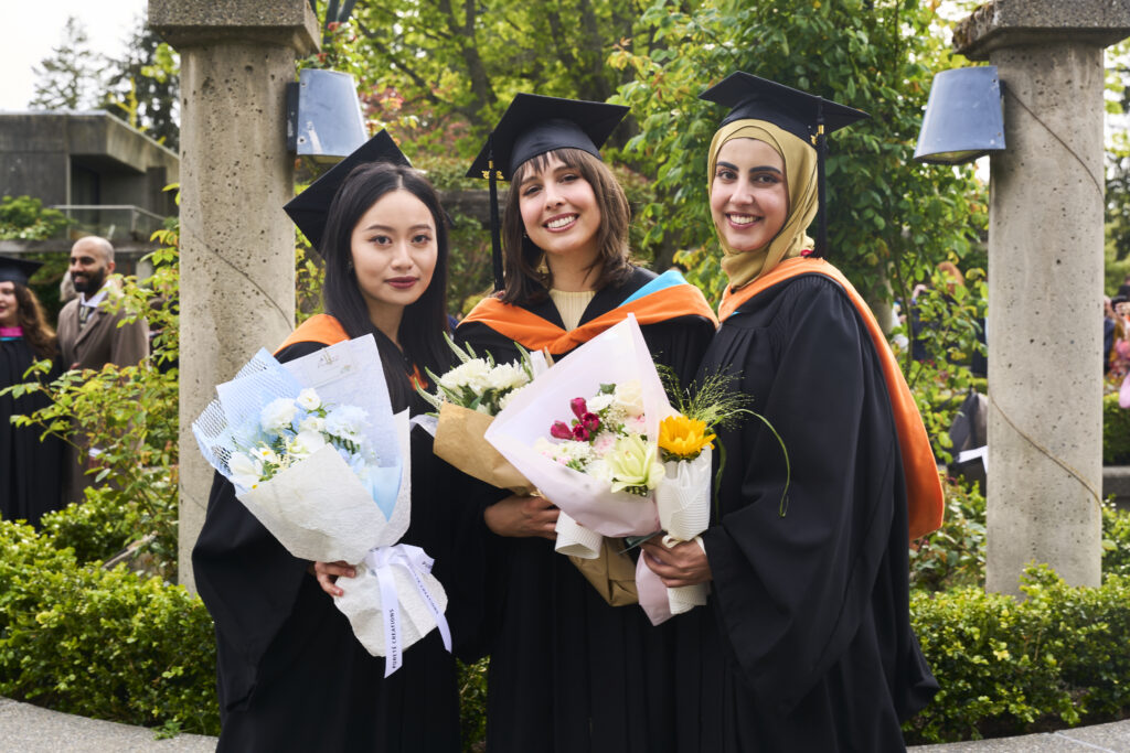 Three graduates in black gowns and orange-trimmed hoods stand together outdoors holding bouquets and smiling at the camera.