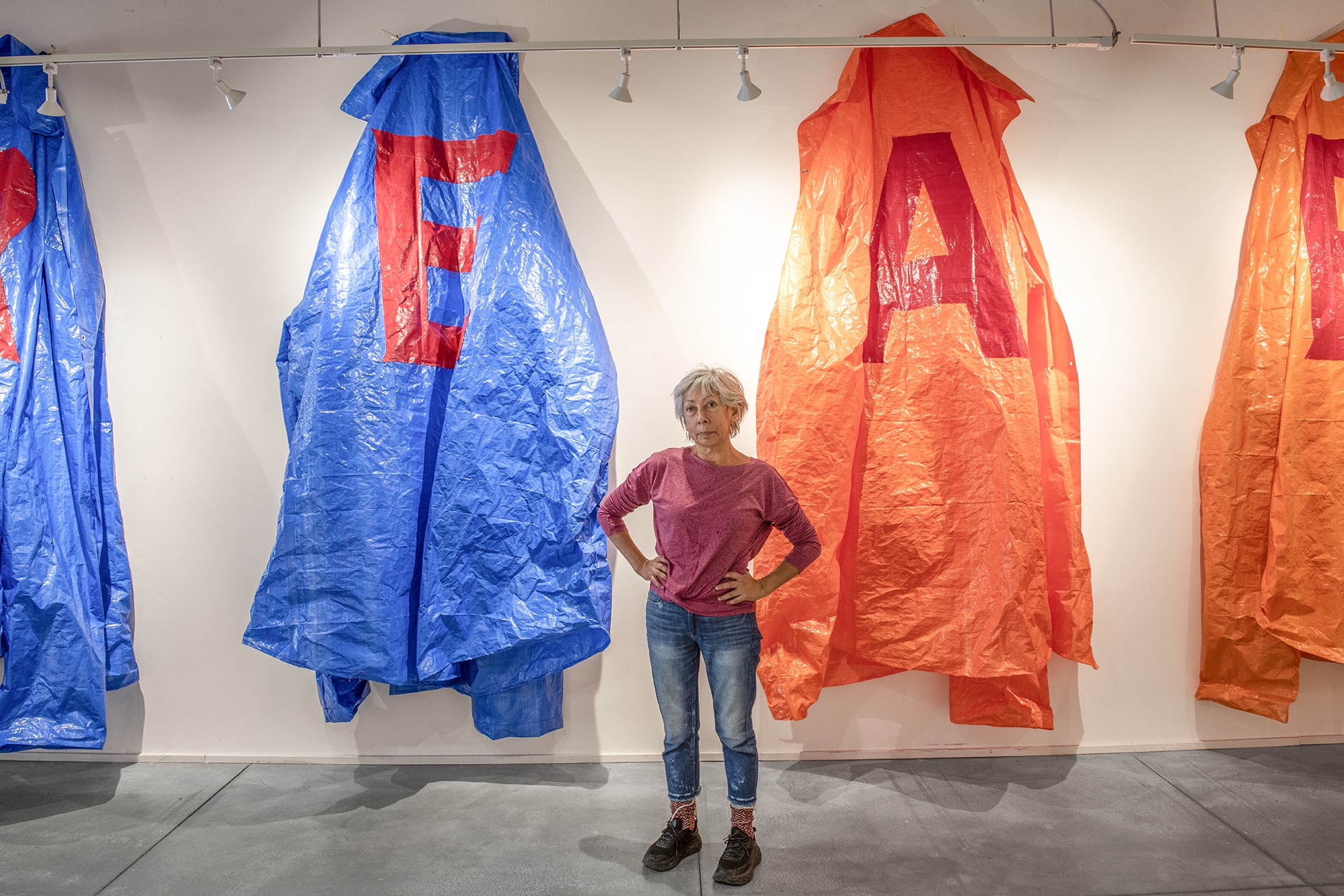 An artist stands in a gallery between large blue and orange hanging garments, their scale towering above as the figure meets the viewer’s gaze.