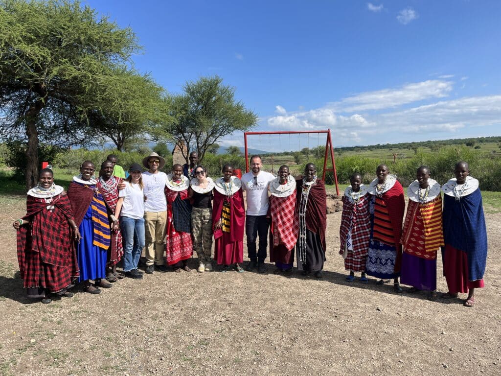 A group of people stand side by side outdoors, facing the camera. Patterned garments in red, blue and white create a bright line across the landscape, with trees, open fields and a swing frame visible behind them under a wide blue sky.