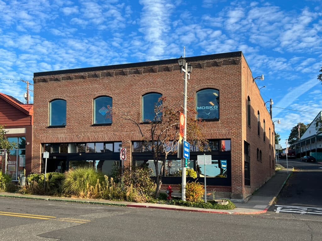 A two-storey brick building sits at a street corner under a bright, cloud-streaked sky. Large windows line the ground floor, with grasses and small trees at the edge of the sidewalk and signage visible in the upper windows.