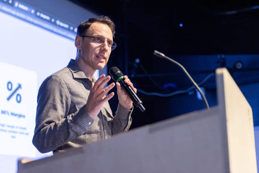 A presenter speaks into a handheld microphone at a podium, one hand raised in emphasis, as a projected slide with a percentage symbol glows behind them in cool blue light.