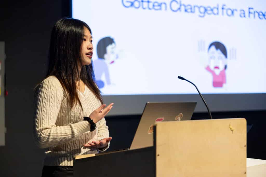 Close-up of a presenter at a podium, gesturing mid-sentence beside an open laptop, with a projected slide and illustrated figures glowing softly in the background.