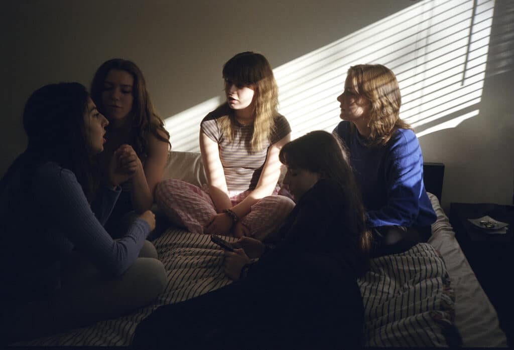 A group of people sit close together on a bed in a softly lit room, engaged in quiet conversation. Sunlight filters through window blinds, casting striped shadows across faces, blankets and the surrounding walls.