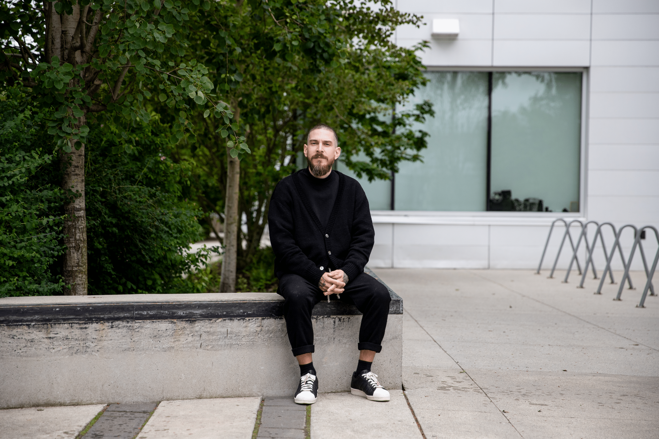 A person sits on a low concrete ledge outdoors, hands loosely clasped, dressed in dark layers, with leafy trees to one side and a modern building behind them, creating a quiet moment of pause within a campus setting.