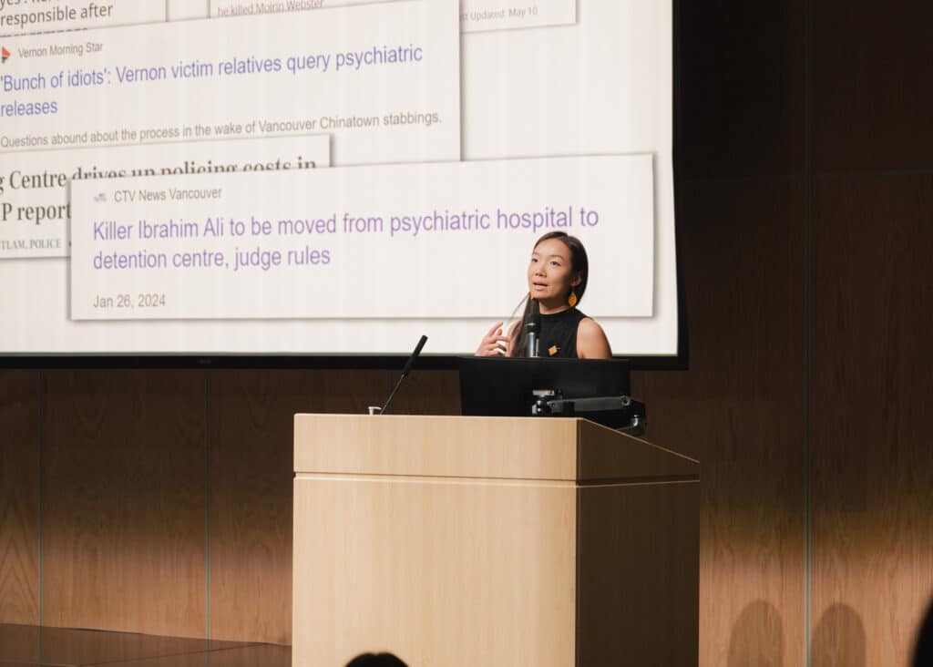 A speaker stands at a podium, gesturing thoughtfully while addressing an audience, as projected news headlines fill the screen behind them.
