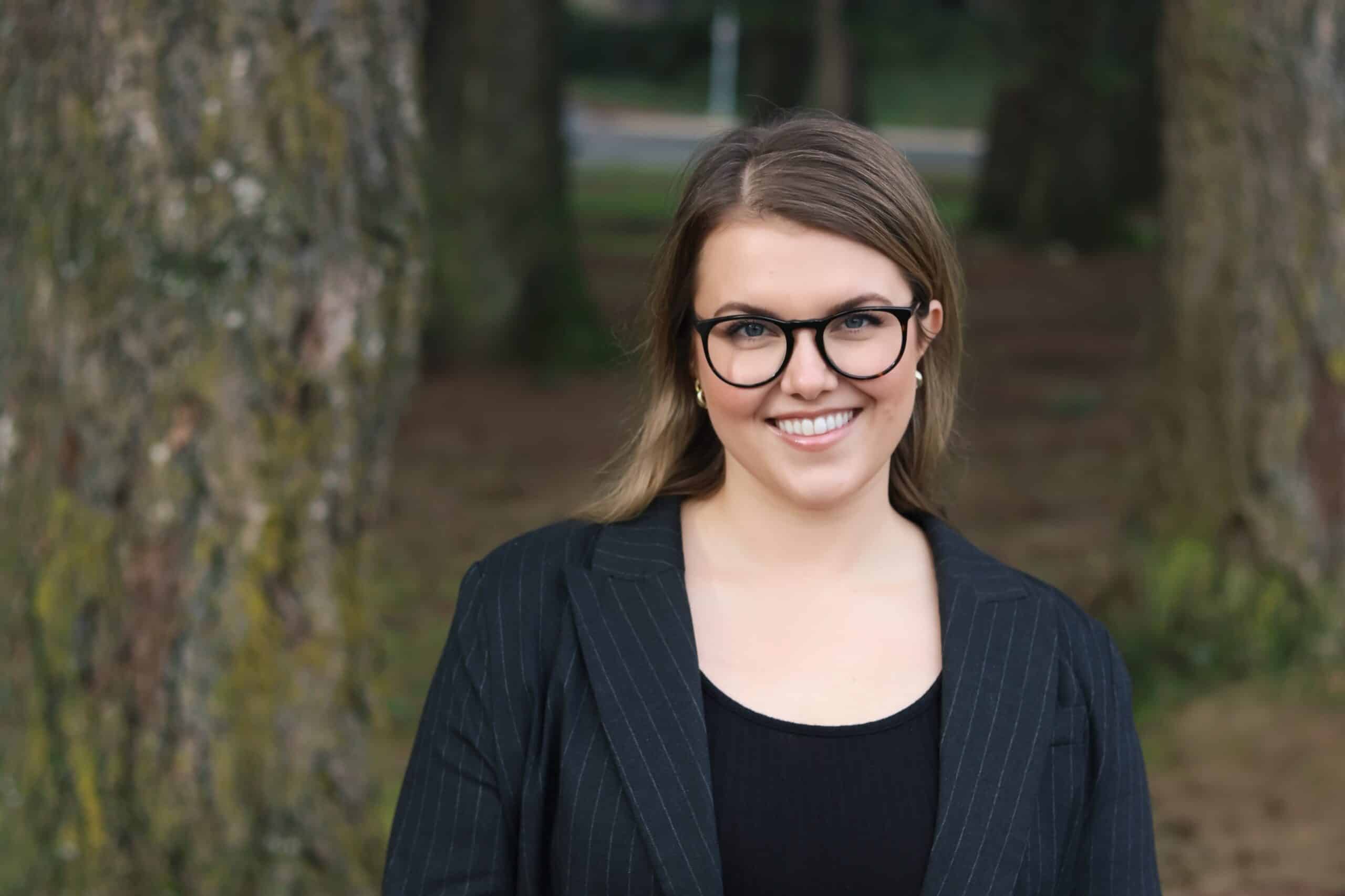 A person wearing glasses and a dark blazer smiles toward the camera while standing outdoors between trees along a path.