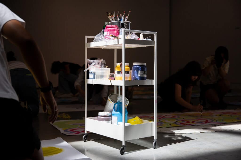 A rolling shelf bearing colourful art supplies including paint and paintbrushes sits on a concrete floor in a beam of sunlight.