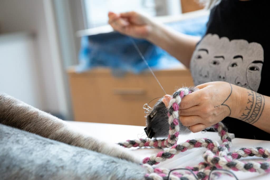 Hands guide a braided strand of wool through a furry form, stitching soft fibres together on a worktable as loose threads and materials gather nearby.